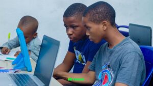 Two young Nigerian boys focused on a laptop during the Netisens ICT Kids Summer Boot Camp 2026 in Uyo, Akwa Ibom, with a third child writing notes in the background